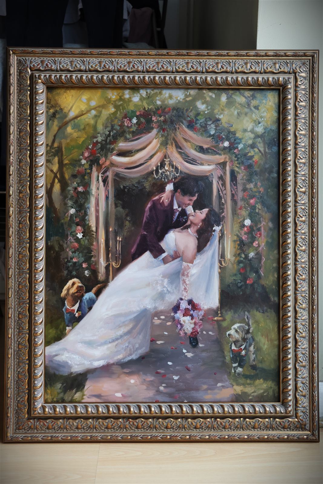 Ornately framed wedding painting of a couple beneath a floral arch, with their two dogs